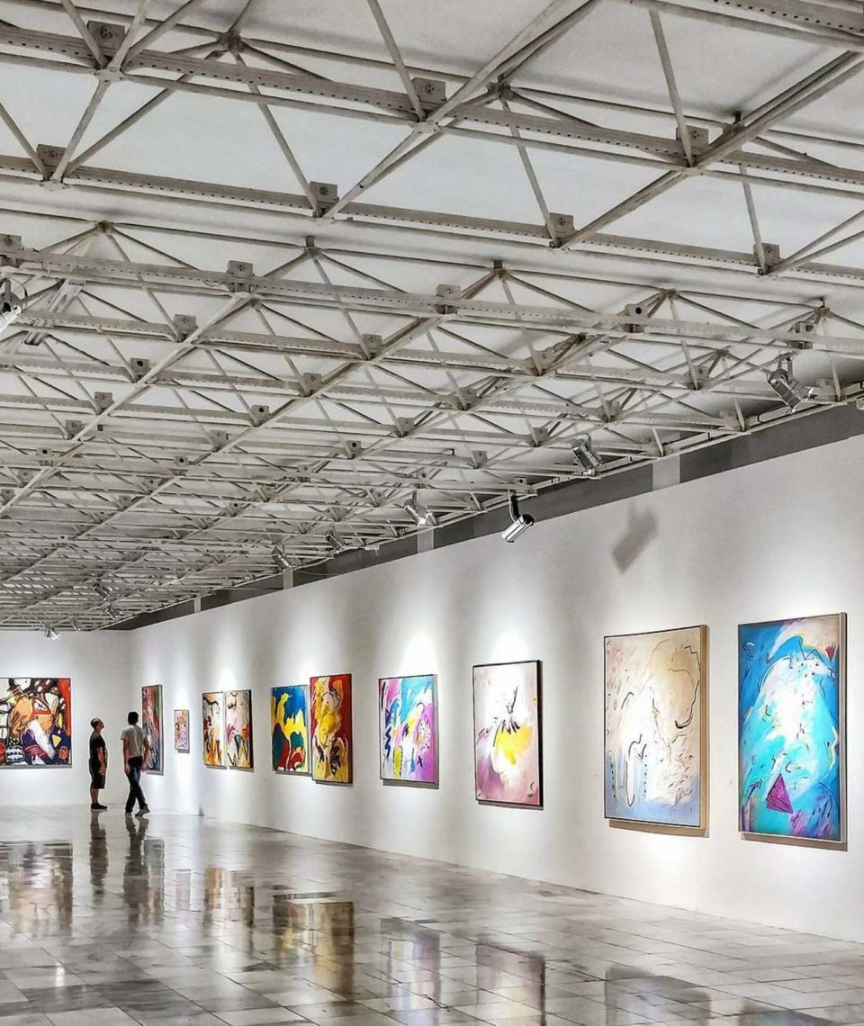 A photo of a gallery space with white walls, and a reflective tile floor, and abstract style paintings on the walls